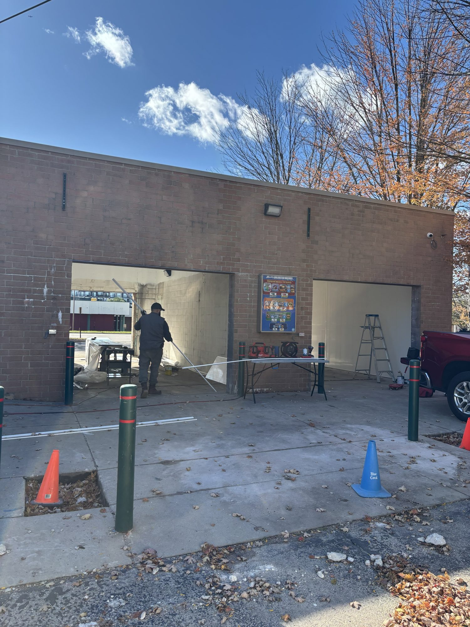 A person paints the interior of a garage-like building with two large open bays; cones, a table, and a ladder are visible outside on a sunny day.