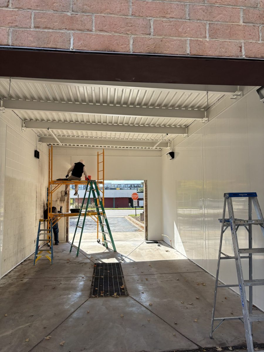 Two people stand on ladders painting the white walls of a car wash bay. Sunlight streams in from the open end, and a stop sign is visible outside.