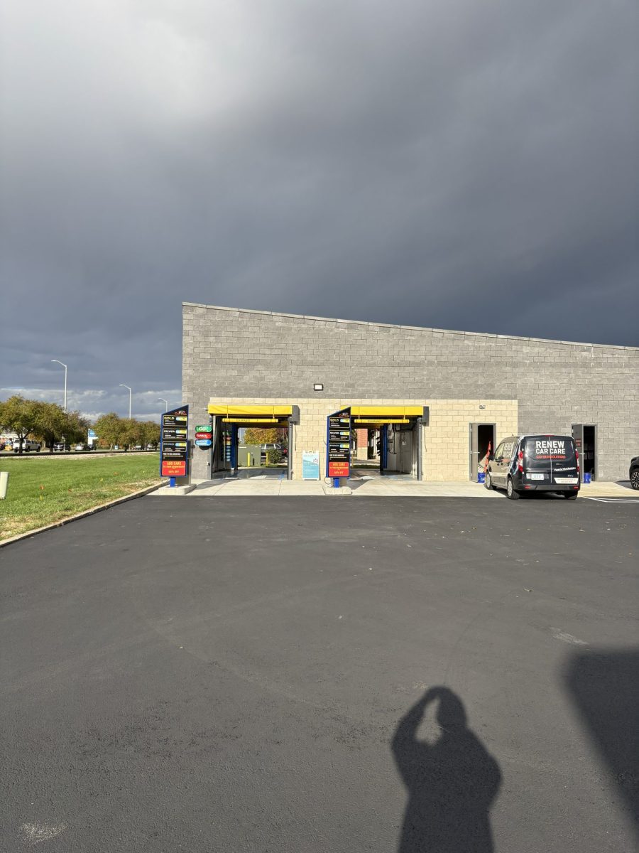Remodel of New Lenox Car Wash with two Petit 360i units now open! A gray, single-story car wash building with yellow awnings and a black van parked on the right, under a cloudy sky. A person's shadow is visible in the foreground on the paved lot.