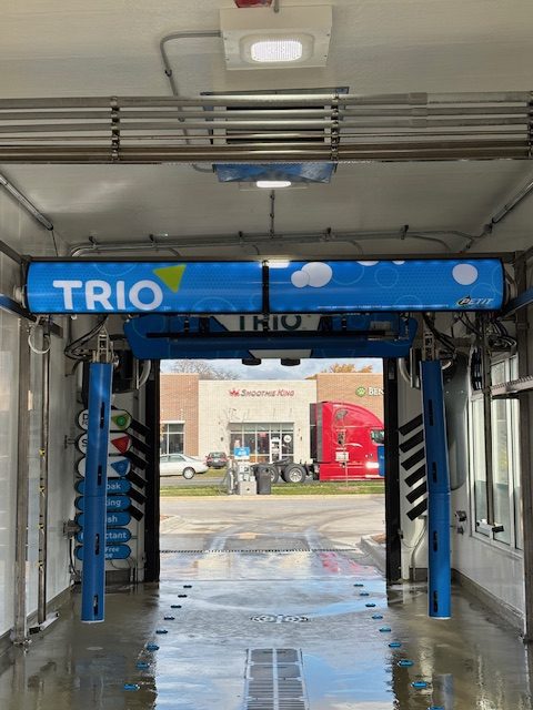 Automated car wash machine labeled "TRIO" inside a bay, with a red truck and buildings visible through the entrance in the background.
