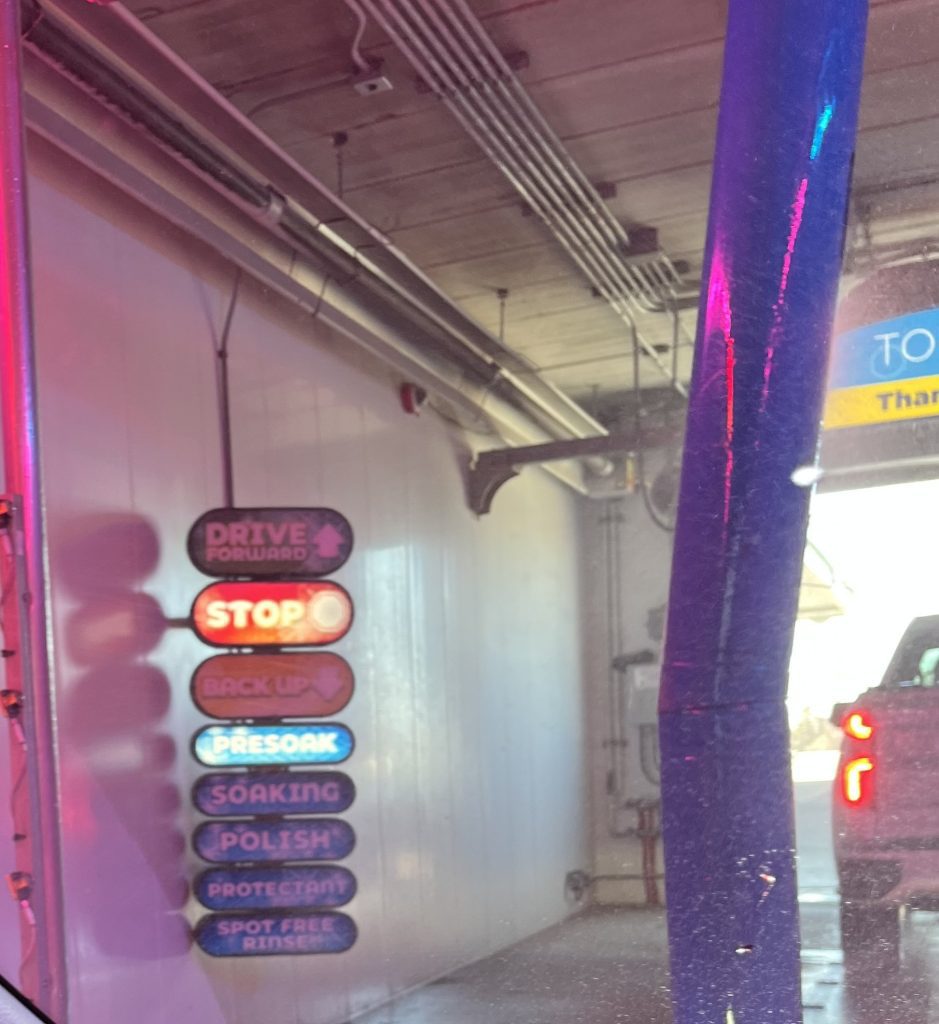 A car approaches the end of an automated car wash tunnel. Various process signs are lit, including stop, presoak, soaking, polish, protectant, and spot free rinse.