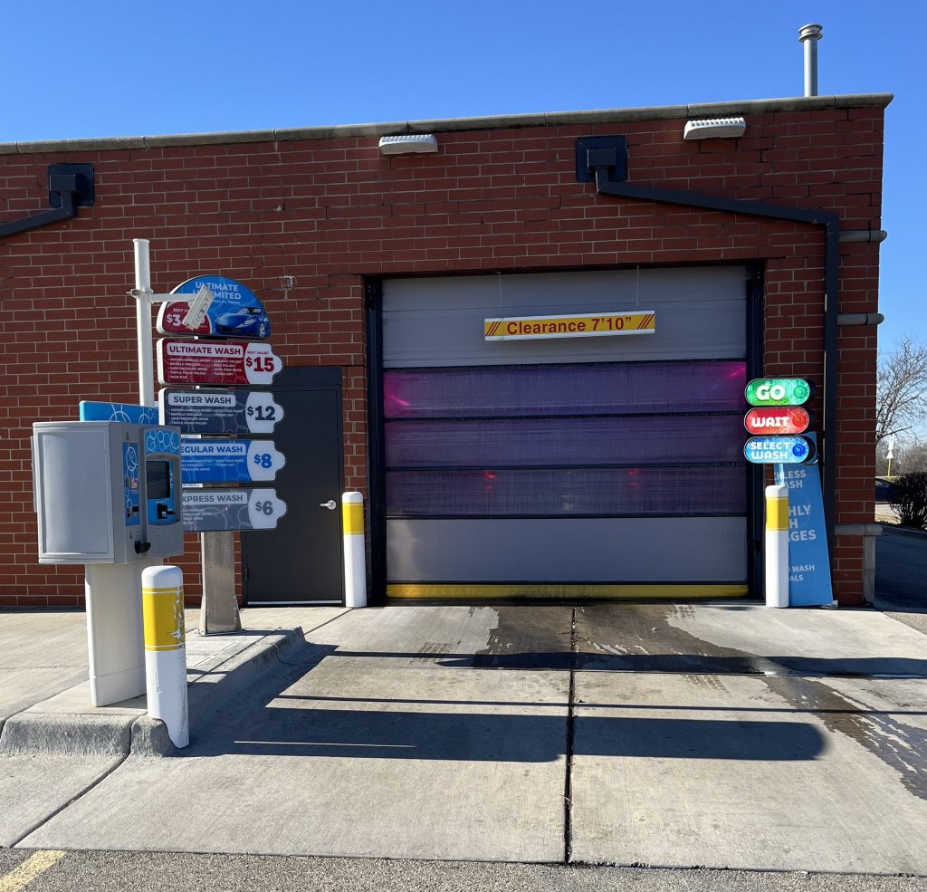 A brick car wash building with a closed garage door, pricing signs, a payment terminal, and clearance height labeled 7'10".