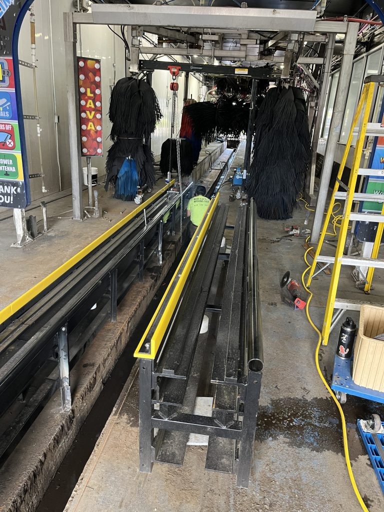 A person in a yellow shirt works inside an automated car wash with brushes and equipment visible; no cars are present on the tracks.