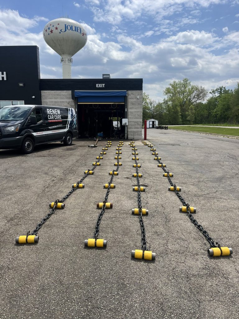 Rows of black chains with yellow rollers lay on the ground outside an auto care garage, with a van parked nearby and a Joliet water tower in the background.