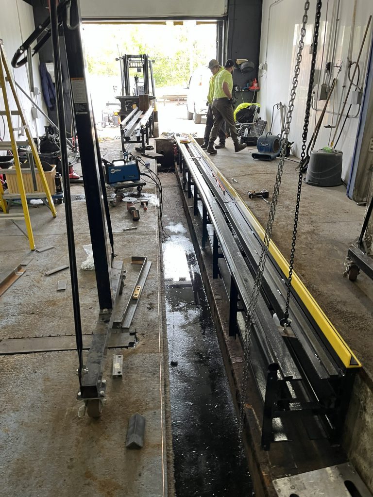 A workshop with long metal beams, tools, and equipment scattered on the floor; a person in a high-visibility shirt works near the open garage door.