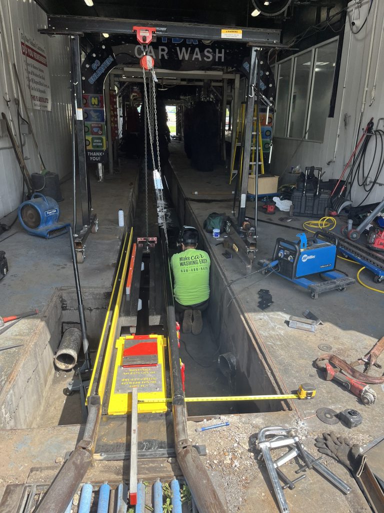 A worker in a green shirt is inside a service pit beneath a car wash conveyor, surrounded by tools and machinery during maintenance or repair work.