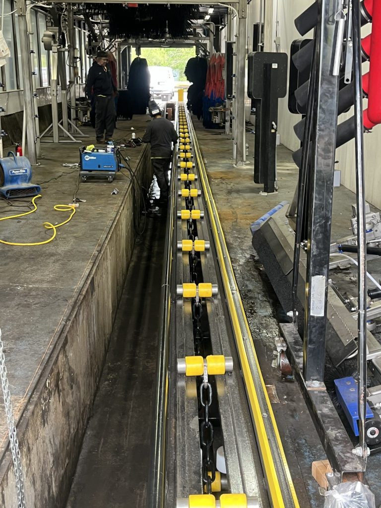A car wash conveyor track under maintenance, with tools, cables, and workers visible along the length of the equipment.