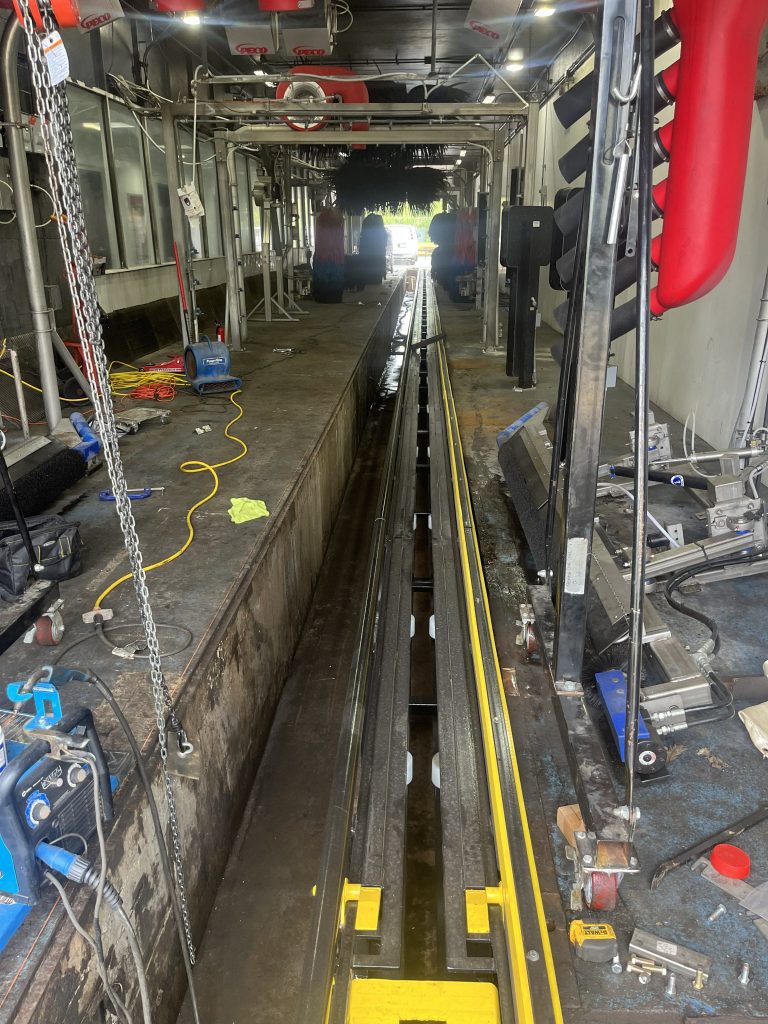 A car wash tunnel with visible tracks, machinery, hoses, and equipment, viewed from the entrance end looking toward the exit.