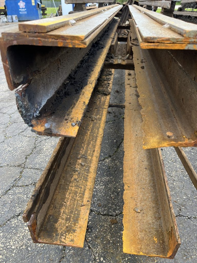 Close-up view of several rusted and weathered steel beams stacked outdoors on cracked pavement.