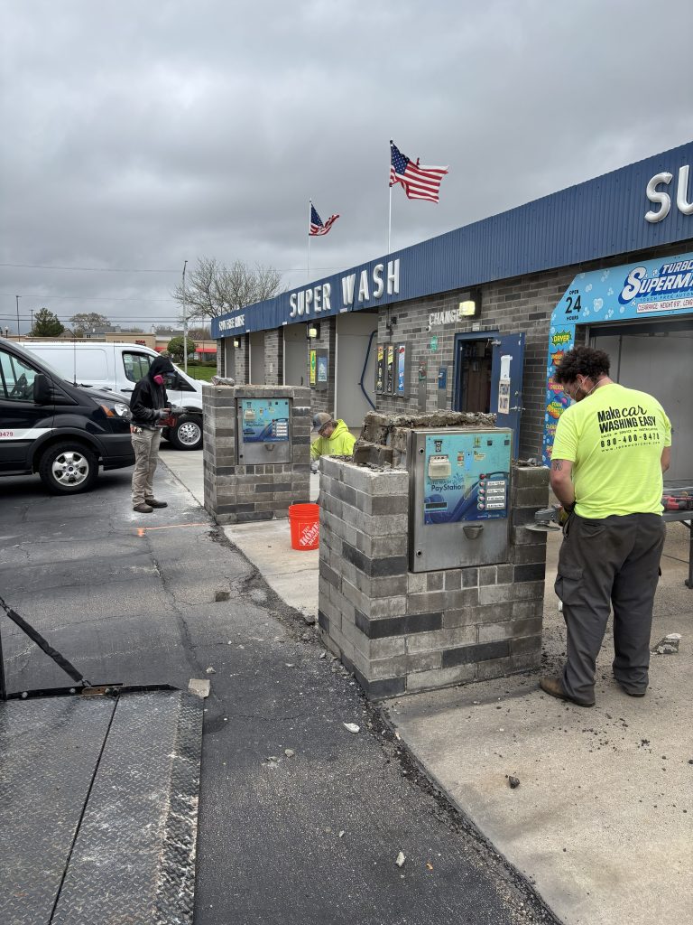 Workers in safety vests repair or construct brick structures near self-service car wash stations outside a Super Wash on a cloudy day. Two American flags are visible on the building.