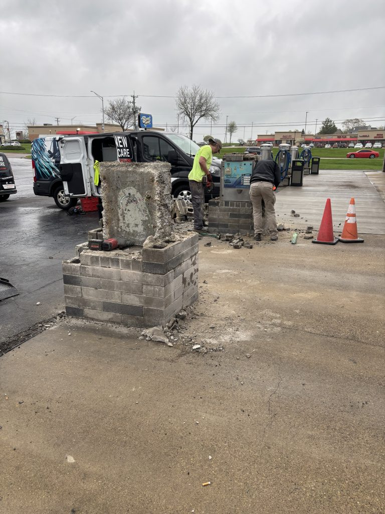 Two workers are demolishing concrete block structures in a parking lot near traffic cones and a work van on a cloudy day.