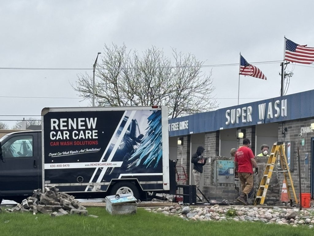 A Renew Car Care van is parked outside a Super Wash building, with workers using ladders and tools near the entrance. Debris and construction materials are visible on the ground.