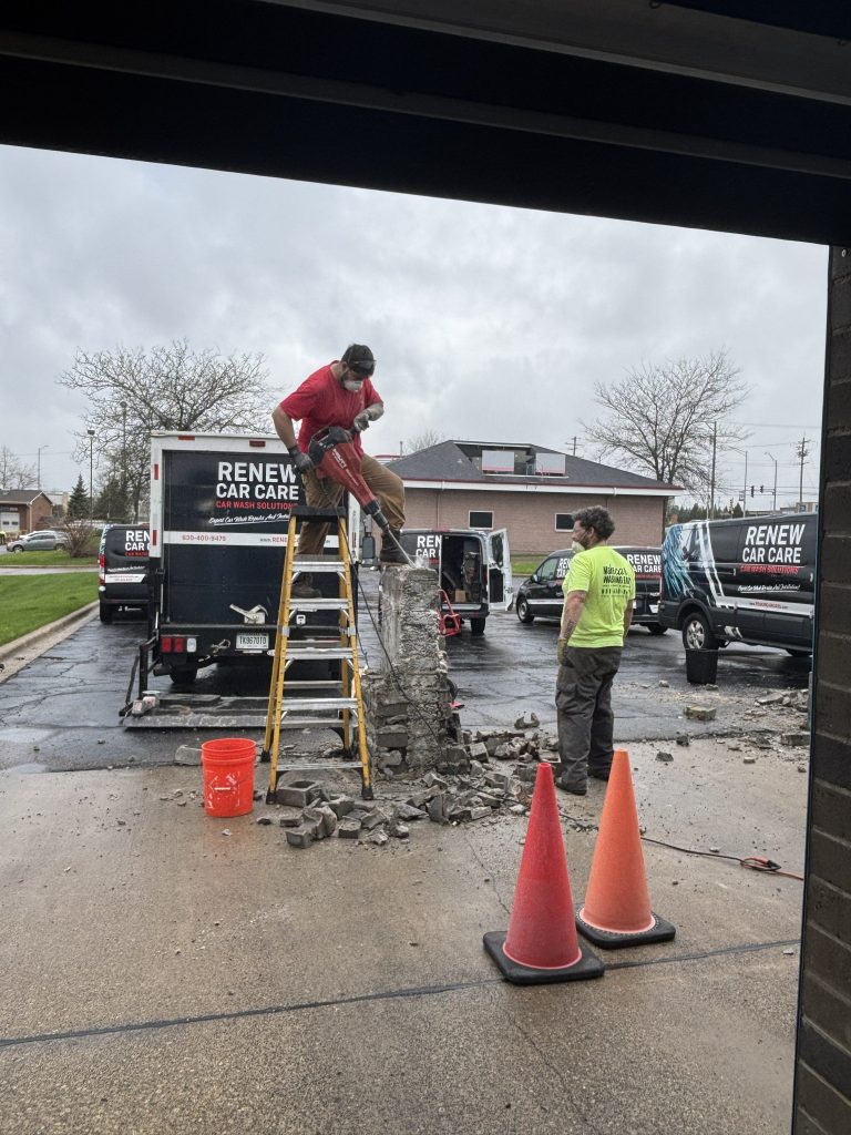 Two workers break concrete outside, one standing on a ladder using a saw and the other observing. Debris covers the ground near safety cones and company vans in the background.