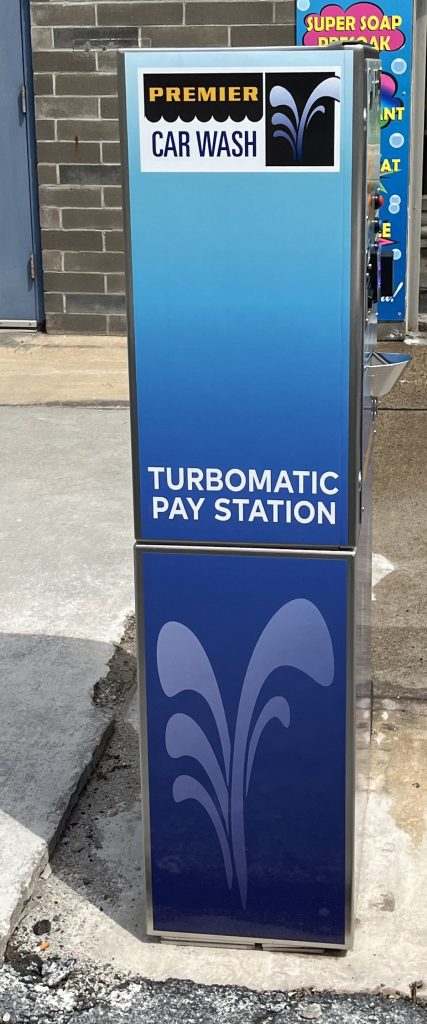 A blue Premier Car Wash Turbomatic Pay Station stands on a concrete surface near a brick wall, with signage and water spray graphics on the machine.