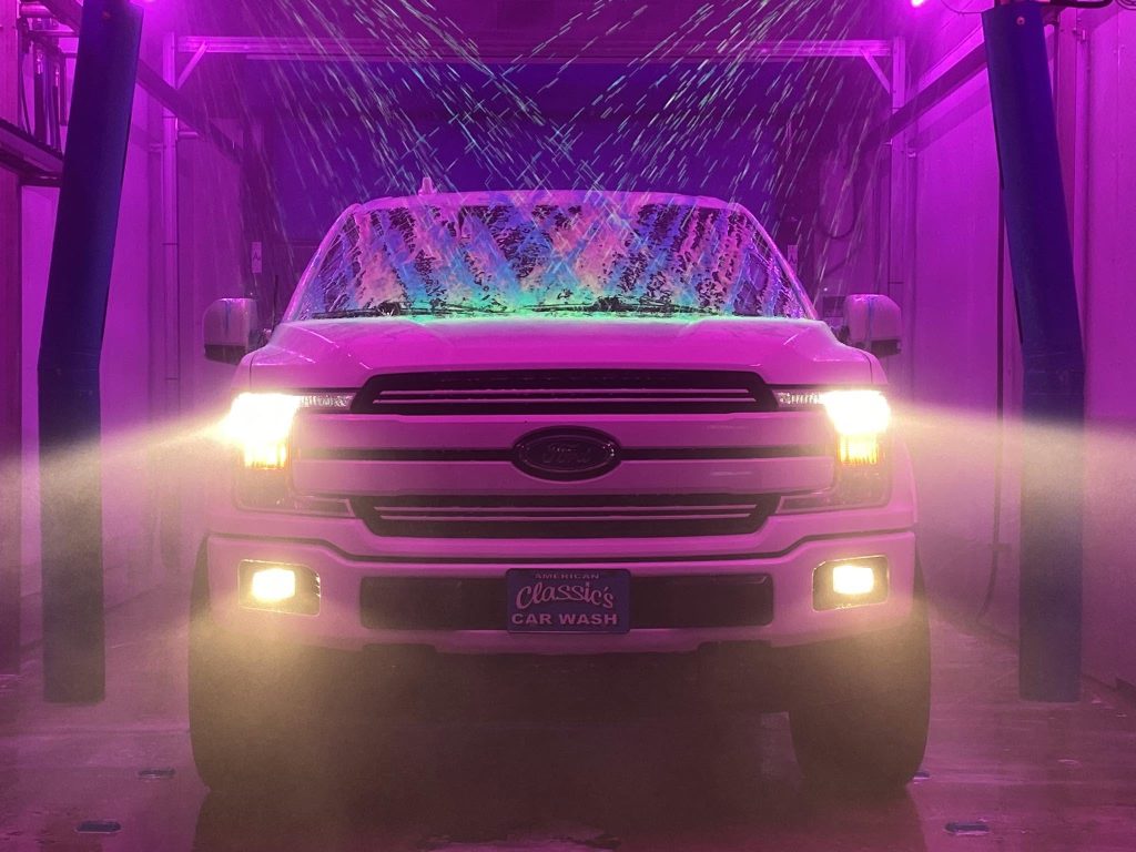 American Classic's Car Wash Finds Success with the Petit Accutrac®360-i A white Ford pickup truck with headlights on moves through a car wash, surrounded by pink and purple lighting and streams of soap and water.