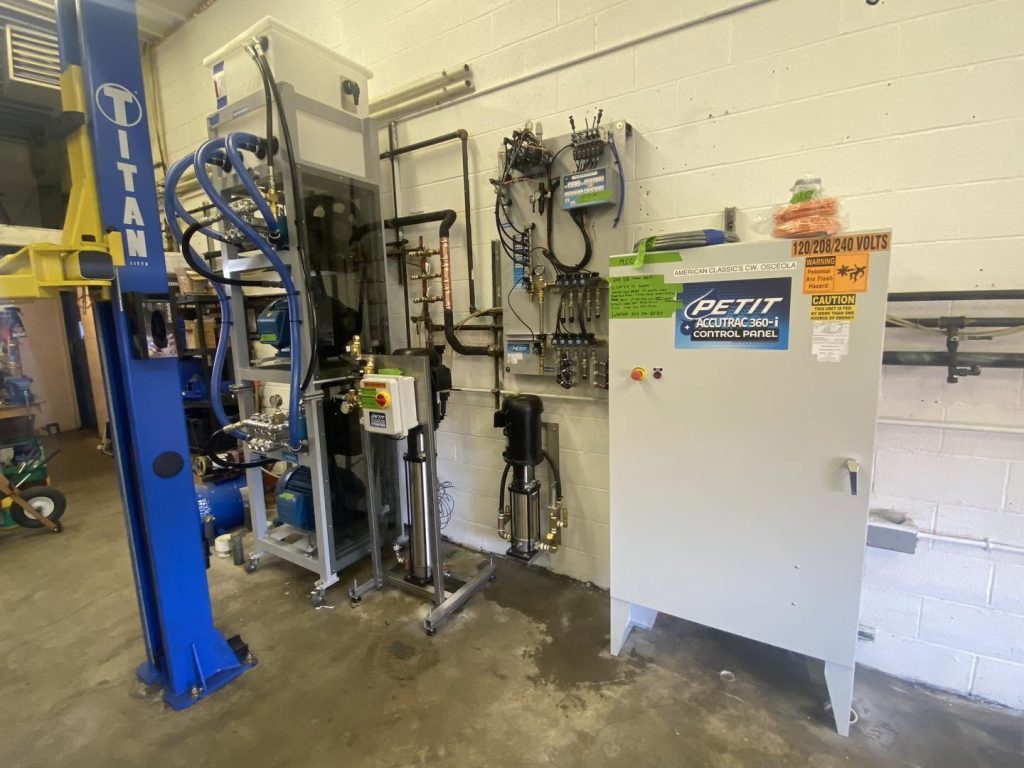 American Classic's Car Wash Finds Success with the Petit Accutrac®360-i Industrial equipment setup in a workshop, featuring a TITAN lift, control panels, pipes, and machinery against a white brick wall.