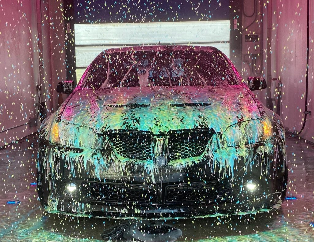 American Classic's Car Wash Finds Success with the Petit Accutrac®360-i A black car is covered in colorful soap suds inside an automatic car wash, with water and foam dripping down the hood and windshield.