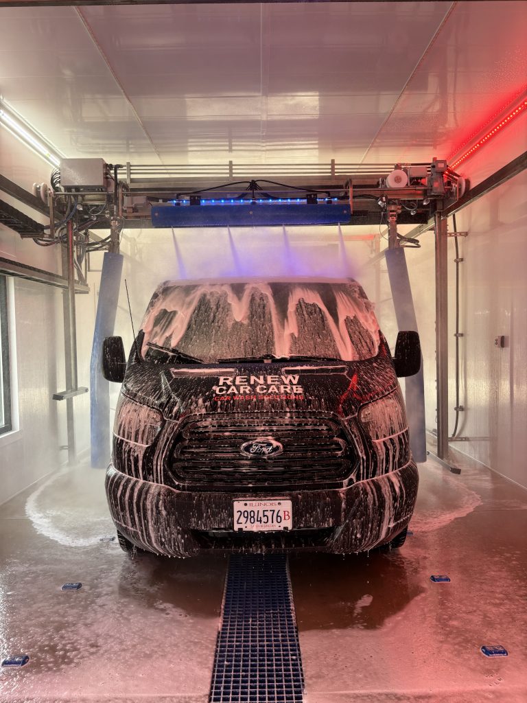 Two New Petit Accutrac® 360-i at Clean Machine Auto Wash A black van covered in white soap foam is parked inside an automatic car wash, with machinery and red and blue lights visible above.