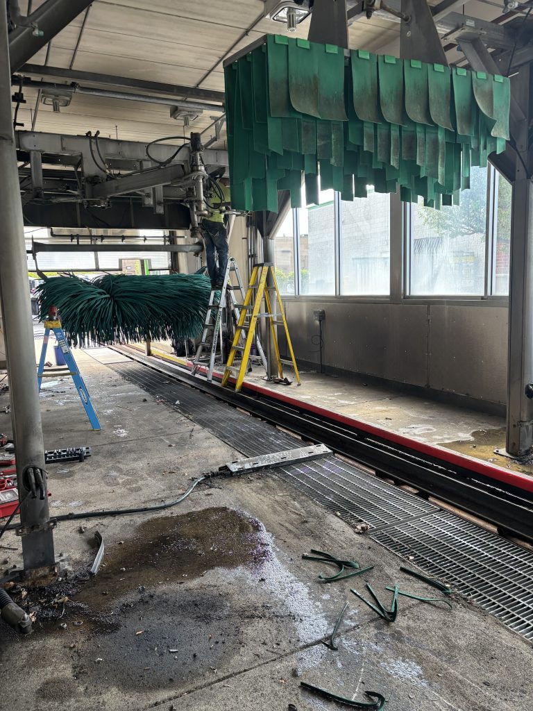 A worker on a stepladder performs maintenance on automatic car wash equipment, including green hanging cleaning strips and a rotating brush.