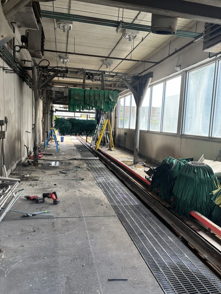 An indoor car wash bay under maintenance, featuring mechanical brushes, a metal floor grate, ladders, and power tools on the concrete ground.